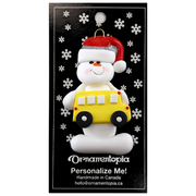Ornament of white snowman in red hat holding a yellow school bus, on display backing card.