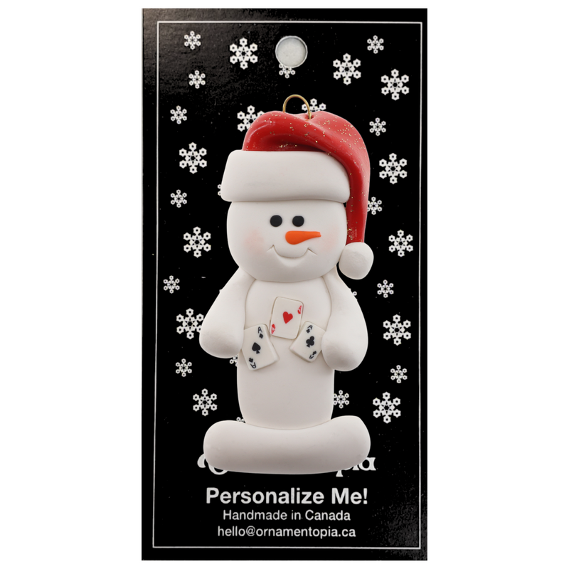 Ornament of white snowman in red hat holding several playing cards, on display backing card.
