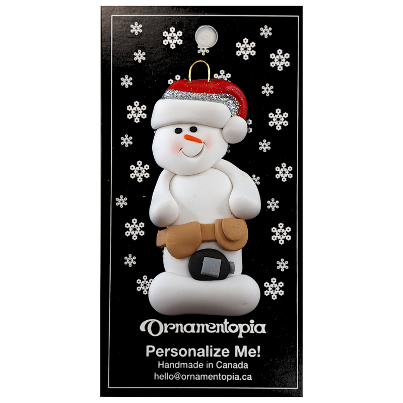 Ornament of white snowman in red hat wearing tool belt with tape measure, on display backing card.