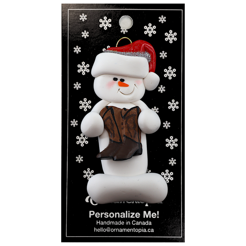 Ornament of white snowman in red hat holding cowboy boots, on display backing card.