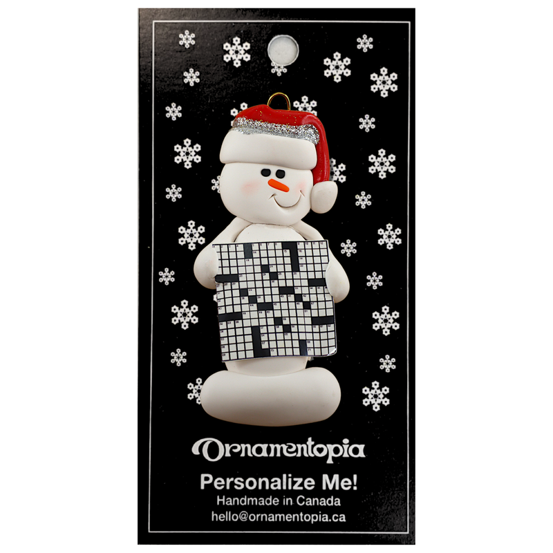 Ornament of white snowman in red hat holding large crossword puzzle, on display backing card.
