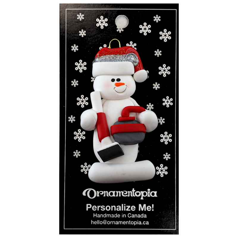 Ornament of white snowman in red hat holding red curling stone and broom, on display backing card.