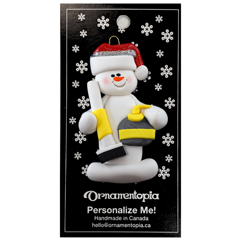 Ornament of white snowman in red hat holding yellow curling stone and broom, on display backing card.