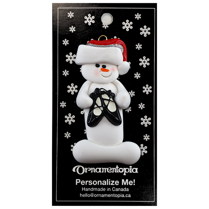 Ornament of white snowman in red hat holding black ballet shoes, on display backing card.
