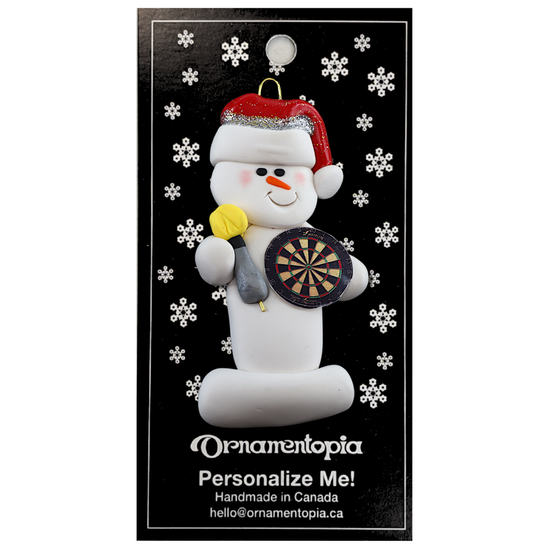 Ornament of white snowman in red hat holding dart board and one dart, on display backing card.