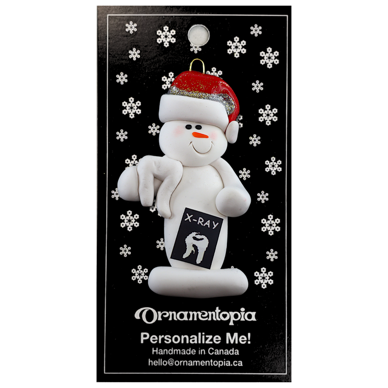 Ornament of white snowman in red hat holding dental x-ray and white tooth, on display backing card.