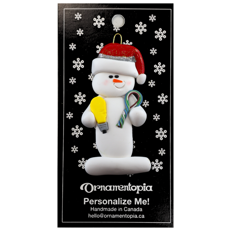 Ornament of white snowman in red hat holding a lightbulb and coil of wire, on display backing card.