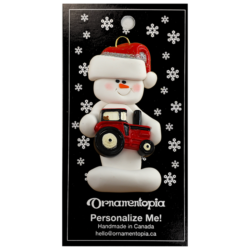 Ornament of white snowman in red hat holding a red tractor, on display backing card.