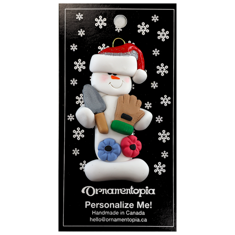 Ornament of white snowman in red hat holding a gardening glove and a shovel with flowers, on display backing card.