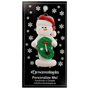 Ornament of white snowman in red hat holding a mini grass green with a golf ball and a flagstick, on display backing card.