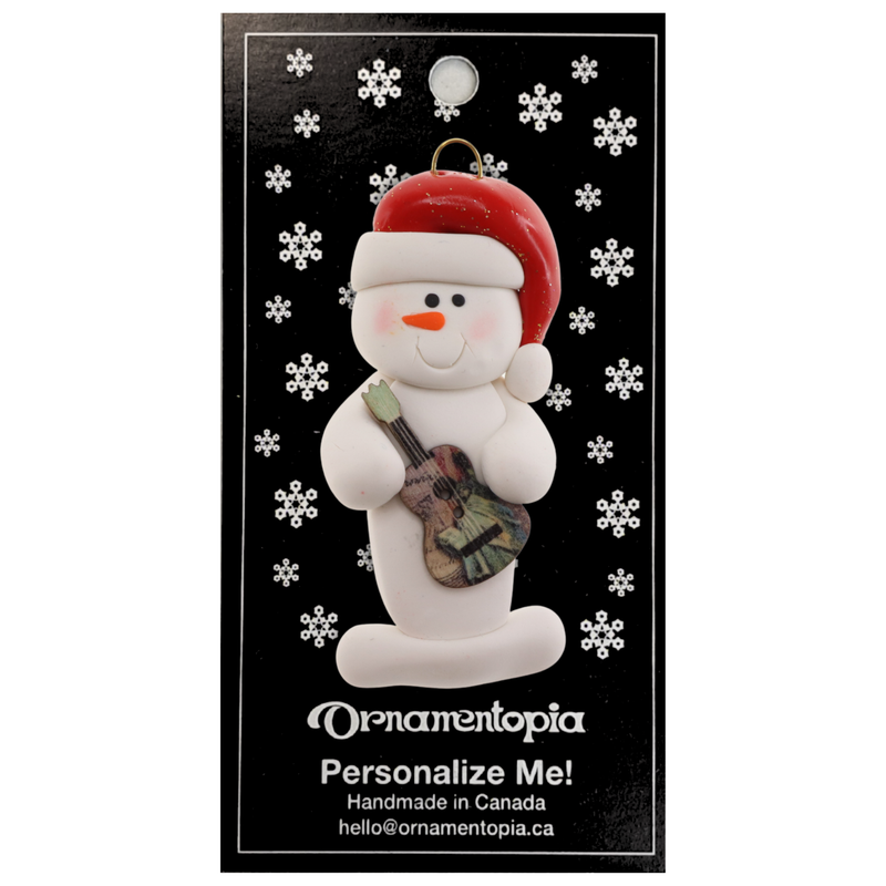 Ornament of white snowman in red hat holding a colourful guitar, on display backing card.