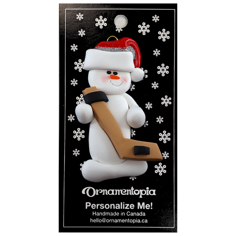 Ornament of white snowman in red hat holding a hockey stick and puck, on display backing card.