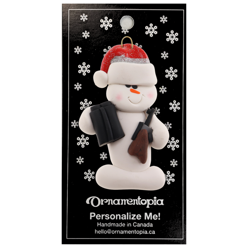 Ornament of white snowman in red hat holding a hunting rifle and pair of binoculars, on display backing card.