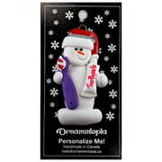 Ornament of white snowman in red hat holding a toothbrush and toothpaste, on display backing card.