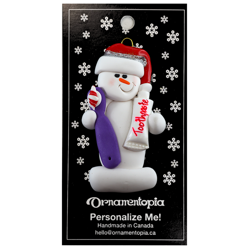 Ornament of white snowman in red hat holding a toothbrush and toothpaste, on display backing card.
