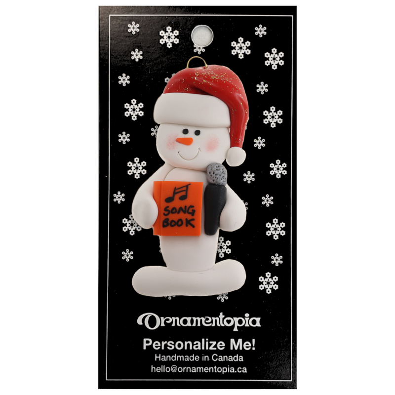 Ornament of white snowman in red hat holding a microphone and orange music book, on display backing card.