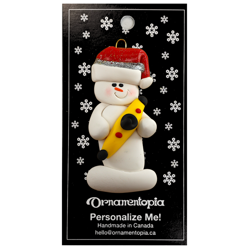 Ornament of white snowman in red hat holding a yellow kayak, on display backing card.