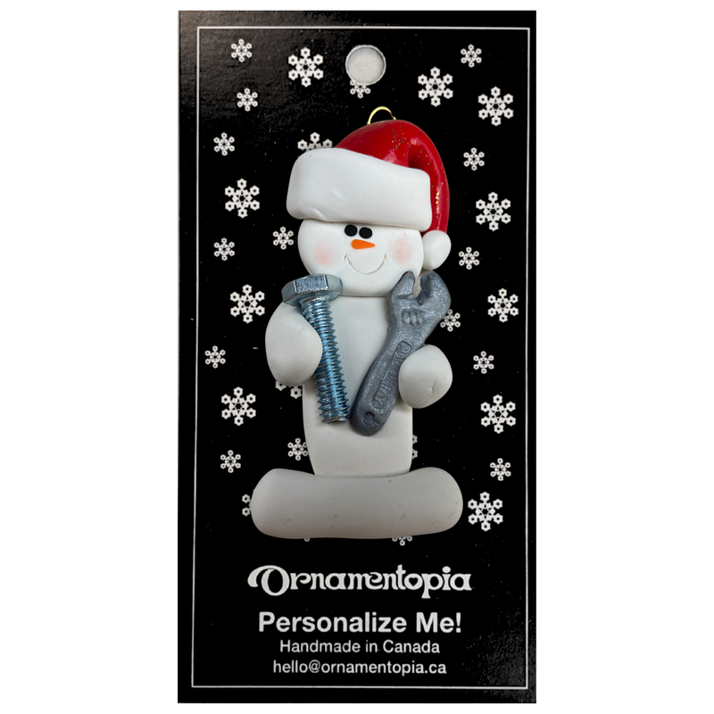 Ornament of white snowman in red hat holding a wrench and a screw, on display backing card.