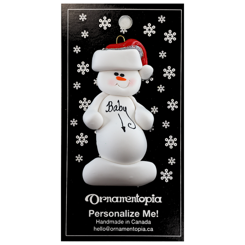 Ornament of white snowman in red hat with a round stomach that says “baby”, on display backing card.