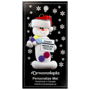 Ornament of white snowman in red hat holding various medications and a medical chart, on display backing card.