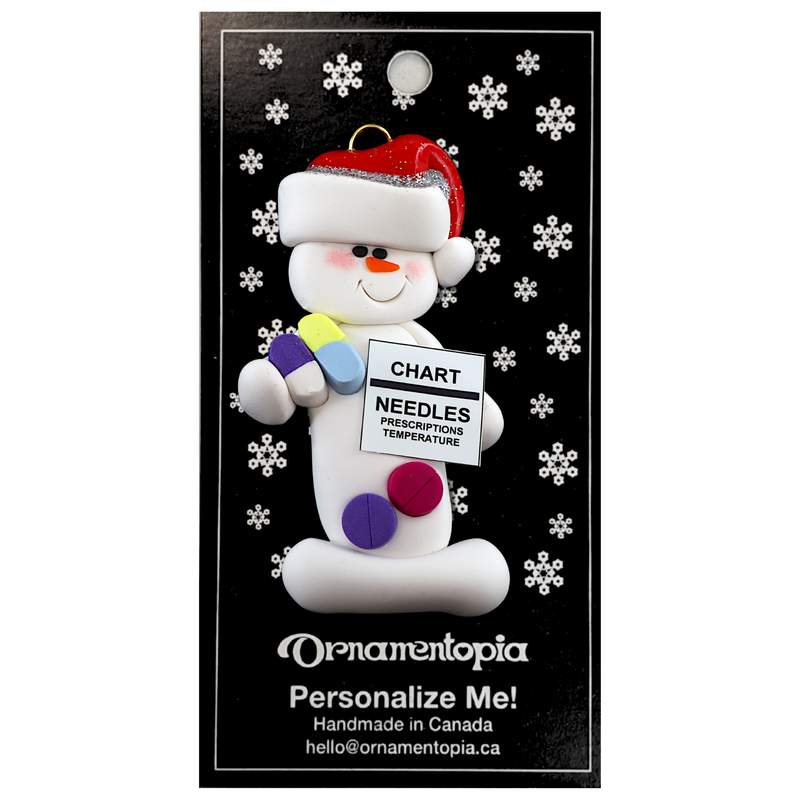 Ornament of white snowman in red hat holding various medications and a medical chart, on display backing card.