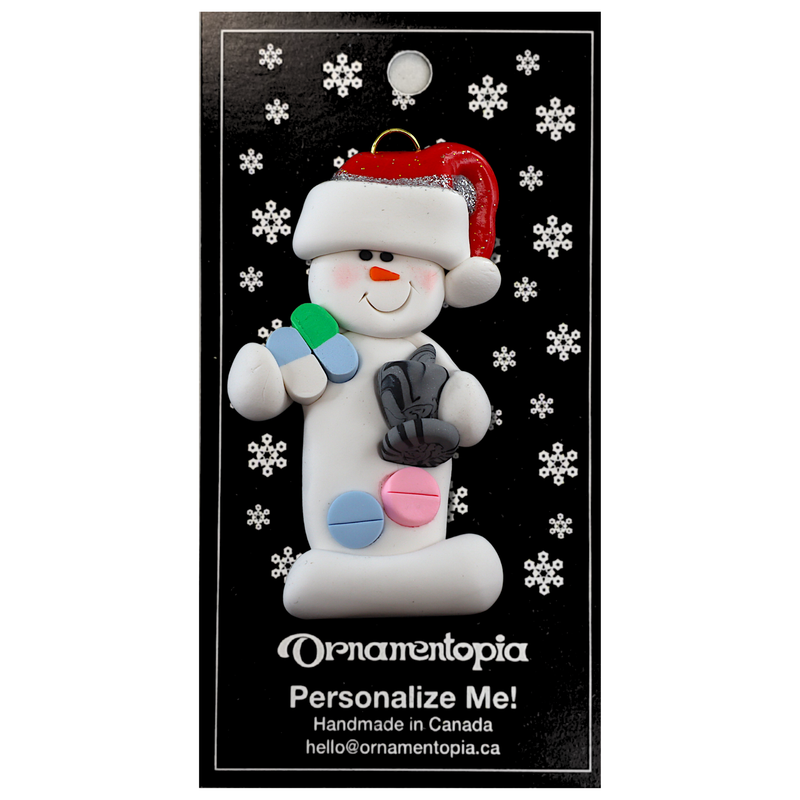 Ornament of white snowman in red hat holding various pills and a mortar and pestle, on display backing card.