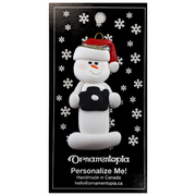 Ornament of white snowman in red hat holding a black camera, on display backing card.