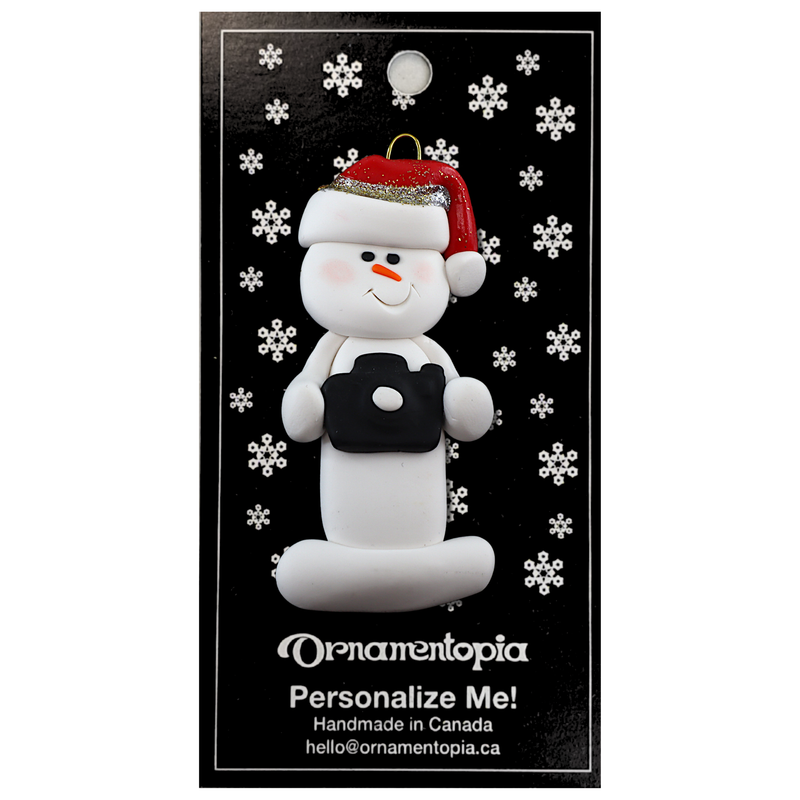 Ornament of white snowman in red hat holding a black camera, on display backing card.