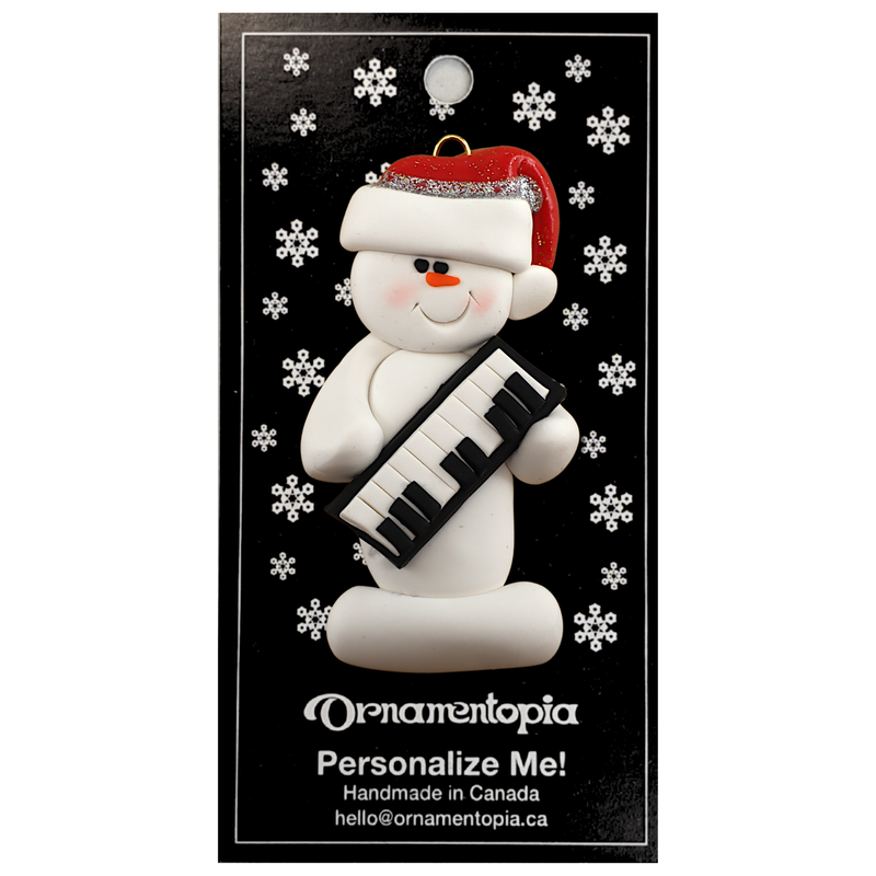 Ornament of white snowman in red hat holding a piano keyboard, on display backing card.
