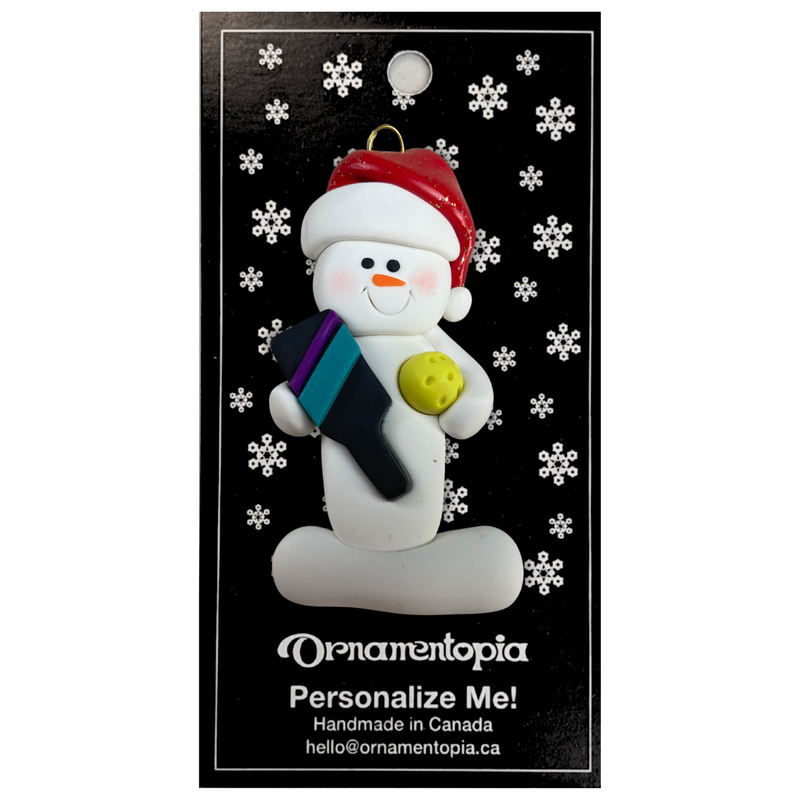 Ornament of white snowman in red hat holding a pickleball paddle and yellow pickleball, on display backing card.