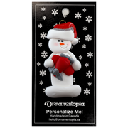 Ornament of white snowman in red hat holding a red airplane, on display backing card.