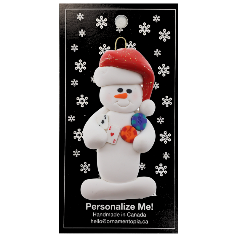 Ornament of white snowman in red hat holding cards and poker chips, on display backing card.