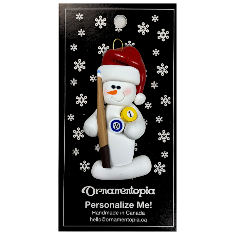 Ornament of white snowman in red hat holding a pool cue and pool balls, on display backing card.