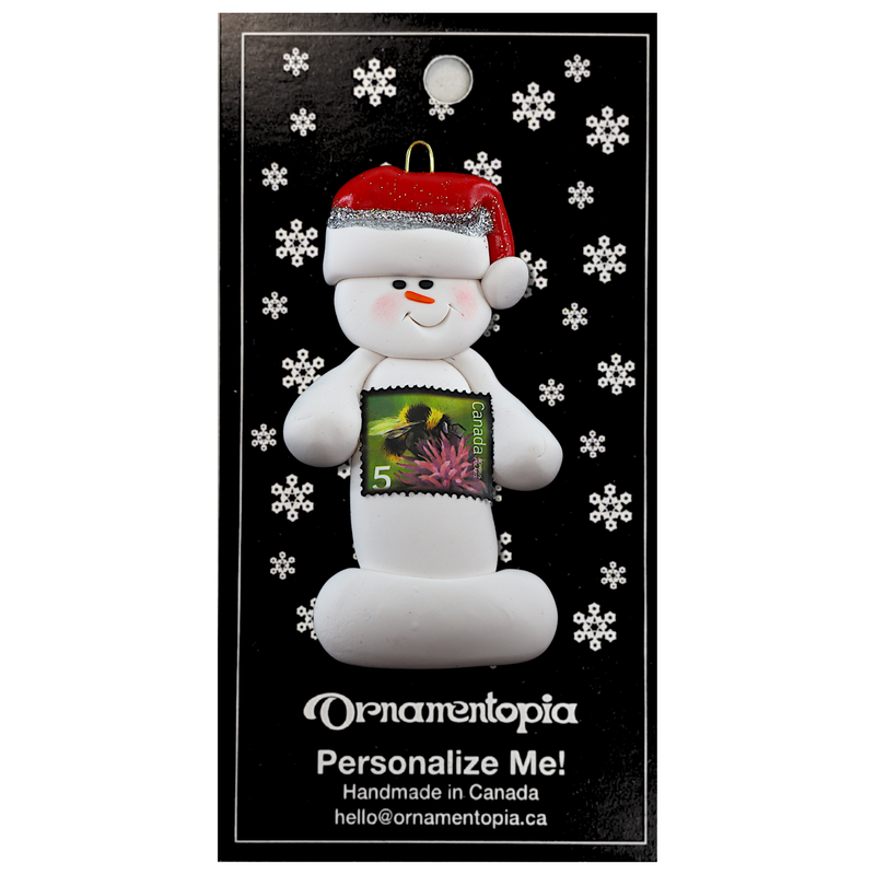 Ornament of white snowman in red hat holding a postal stamp, on display backing card.