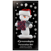 Ornament of white snowman in red hat holding a quilt block and pair of scissors, on display backing card.