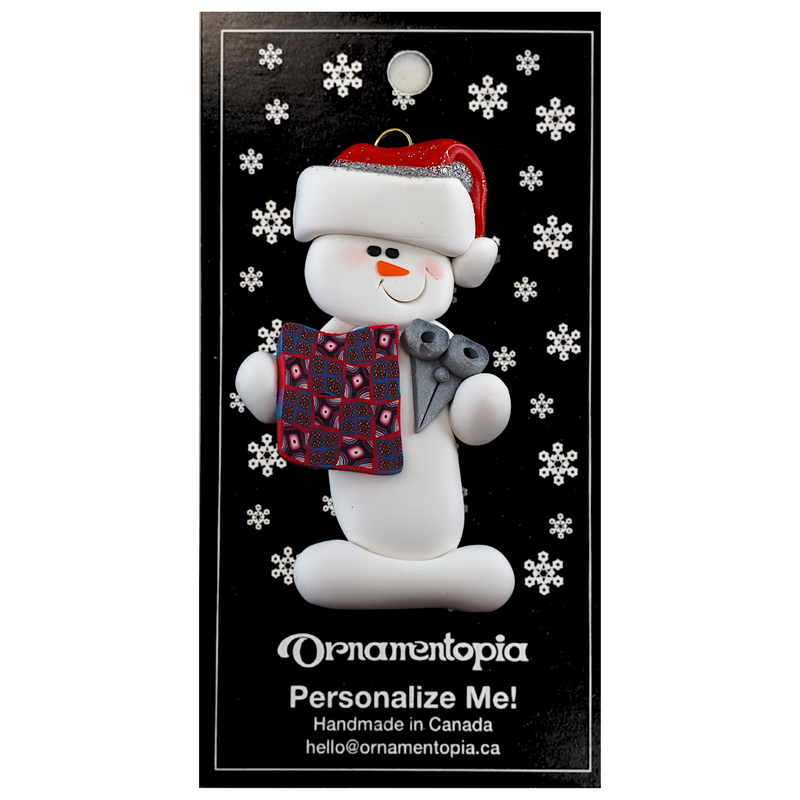 Ornament of white snowman in red hat holding a quilt block and pair of scissors, on display backing card.