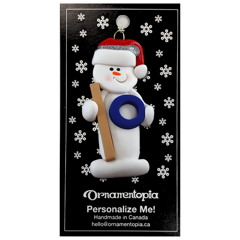 Ornament of white snowman in red hat holding a ringette stick and blue ring, on display backing card.