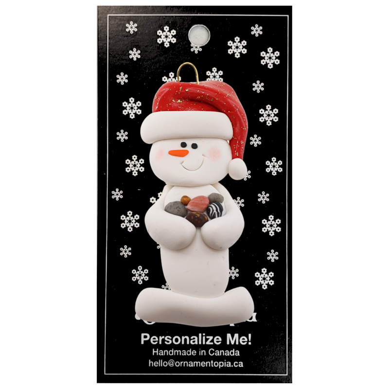 Ornament of white snowman in red hat holding several small rocks, on display backing card.