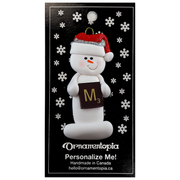Ornament of white snowman in red hat holding a scrabble tile, on display backing card.