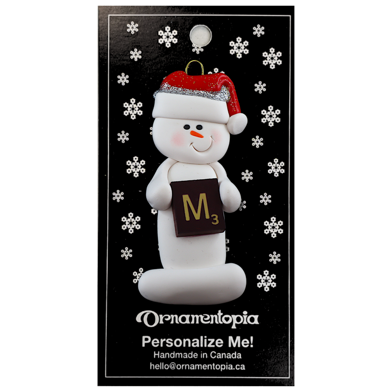 Ornament of white snowman in red hat holding a scrabble tile, on display backing card.