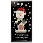 Ornament of white snowman in red hat holding a pair of scissors and various scrapbooking papers, on display backing card.