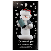 Ornament of white snowman in red hat holding a spool of thread and scissors, on display backing card.