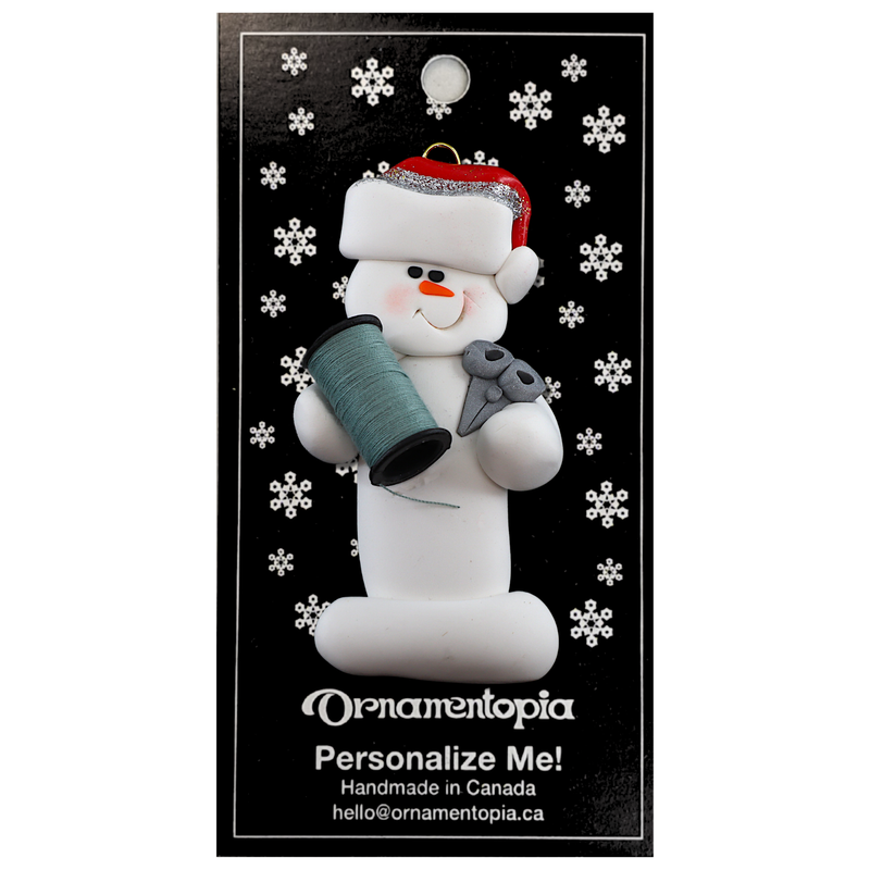 Ornament of white snowman in red hat holding a spool of thread and scissors, on display backing card.