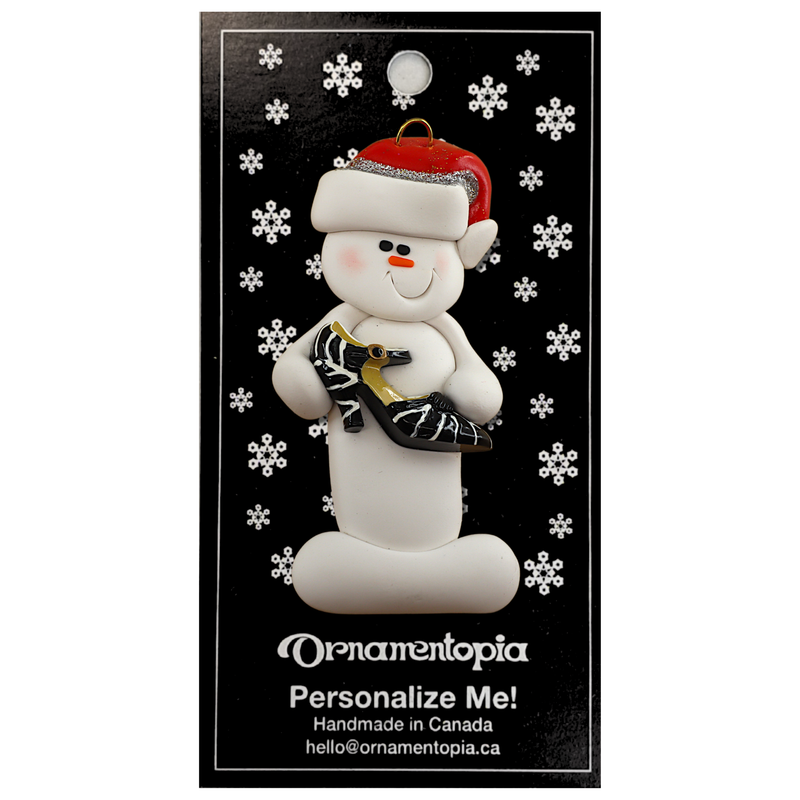 Ornament of white snowman in red hat holding a fancy shoe, on display backing card.