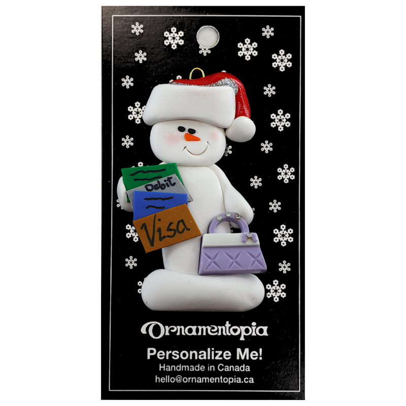 Ornament of white snowman in red hat holding a purse and various bank cards, on display backing card.