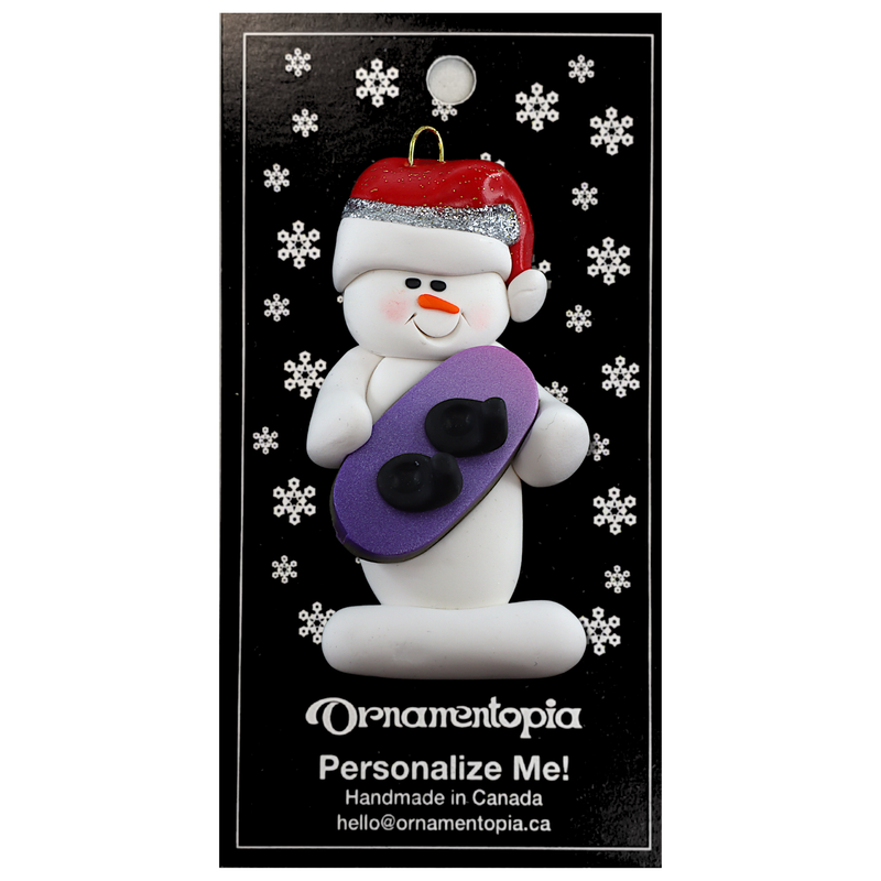 Ornament of white snowman in red hat holding a vibrant snowboard, on display backing card.
