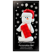 Ornament of white snowman in red hat holding a plastic building brick, on display backing card.