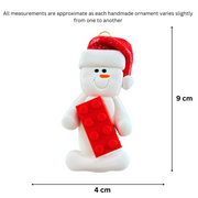 Ornament of white snowman in red hat holding a plastic building brick, graphed to show size (4x9cm).
