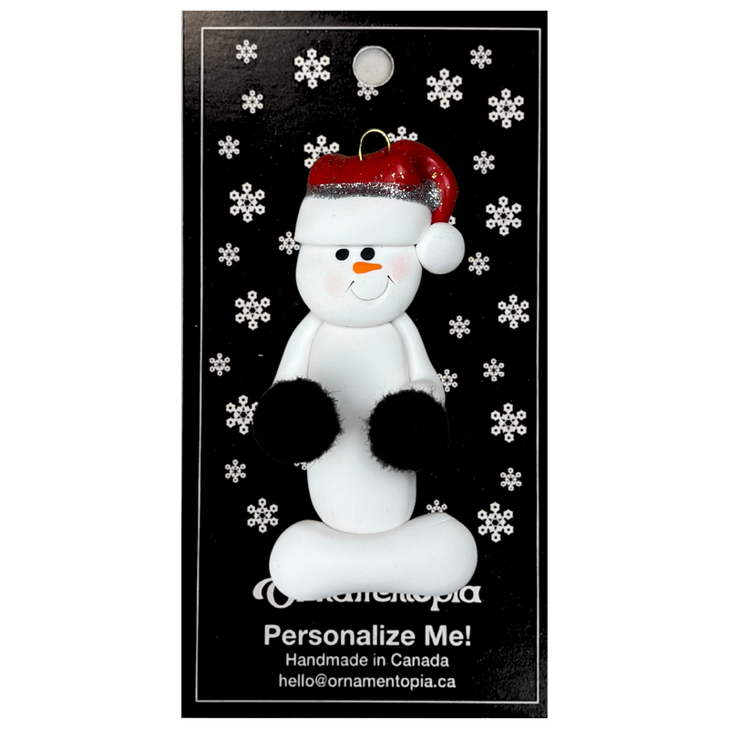 Ornament of white snowman in red hat holding black cheerleader pom-poms, on display backing card.