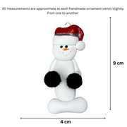 Ornament of white snowman in red hat holding black cheerleader pom-poms, graphed to show size (4x9cm).